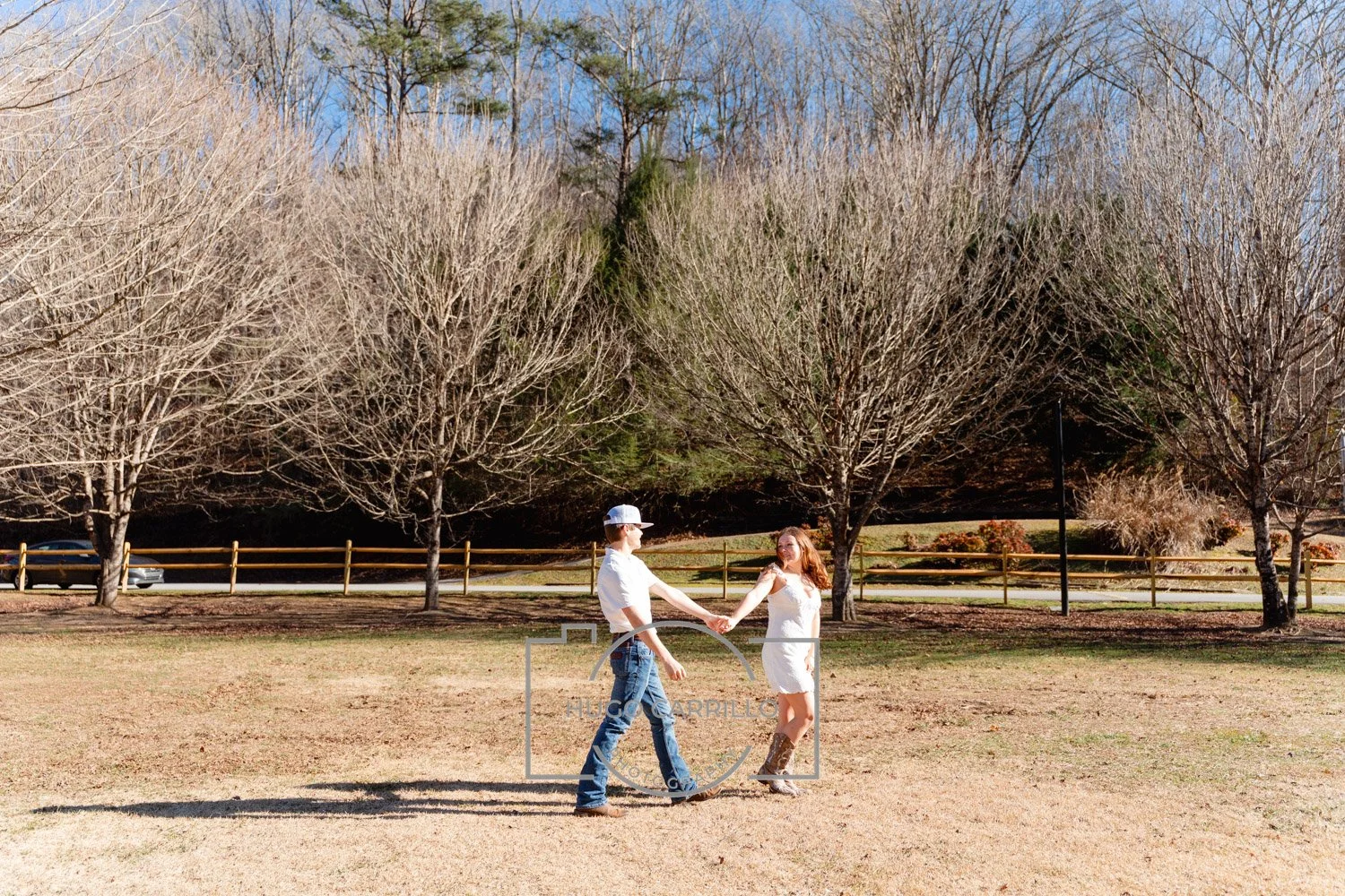 A couple holding hands, dancing in a park with leafless trees and a wooden fence in the background on a sunny day.