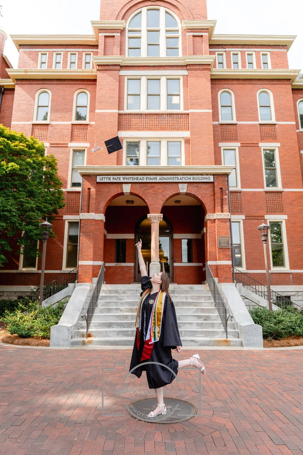 A young woman in a graduation gown and cap throws her cap into the air in front of a brick university building called Lettie Pate Whitehead Evans Administration Building. She stands on a manhole cover and poses with one arm raised.