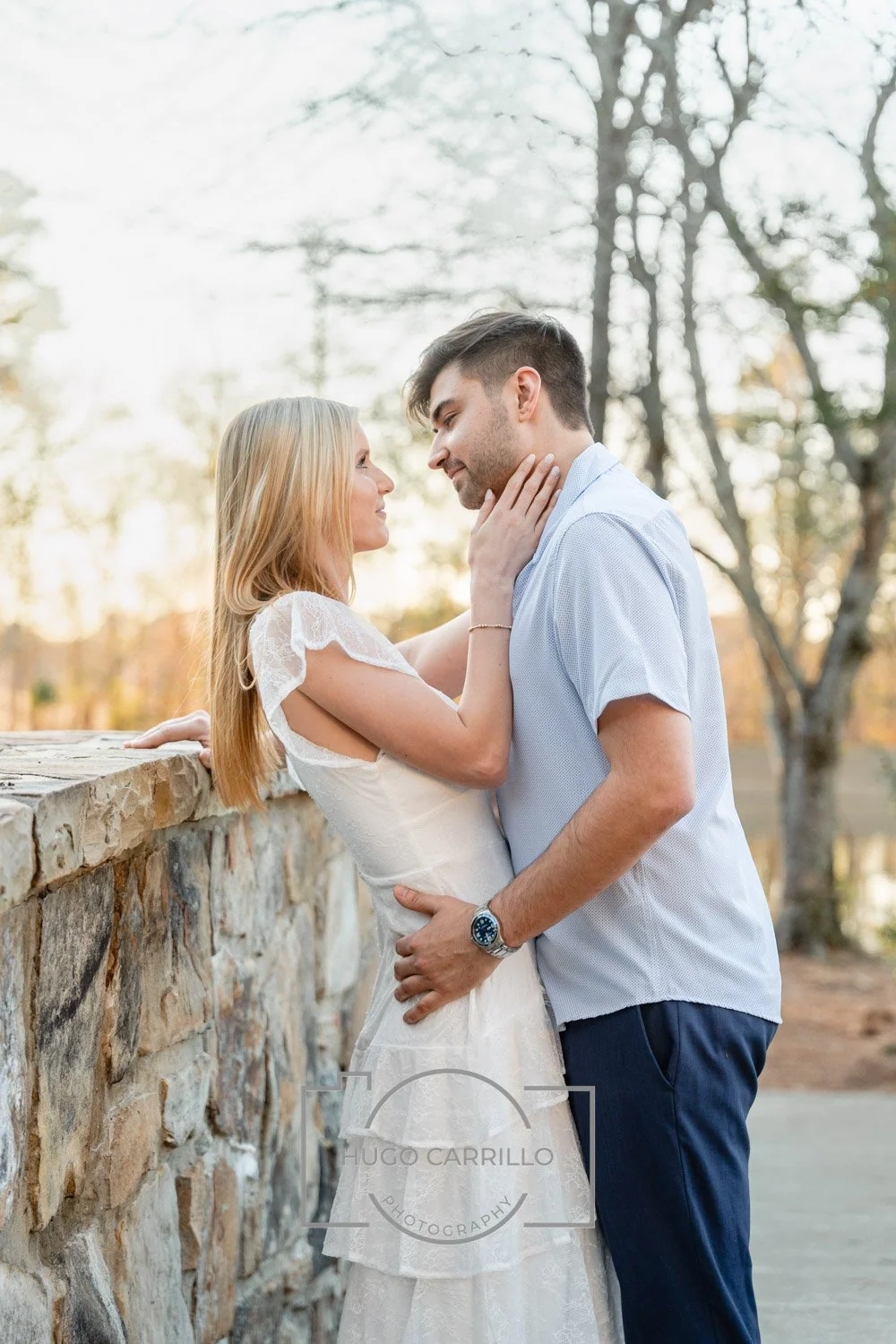 A couple standing close to each other outdoors during sunset, with the woman touching the man's face, both gazing into each other's eyes.