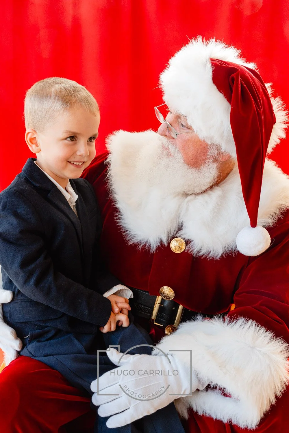 A young boy sitting on Santa Claus's lap, both smiling, with a red background.