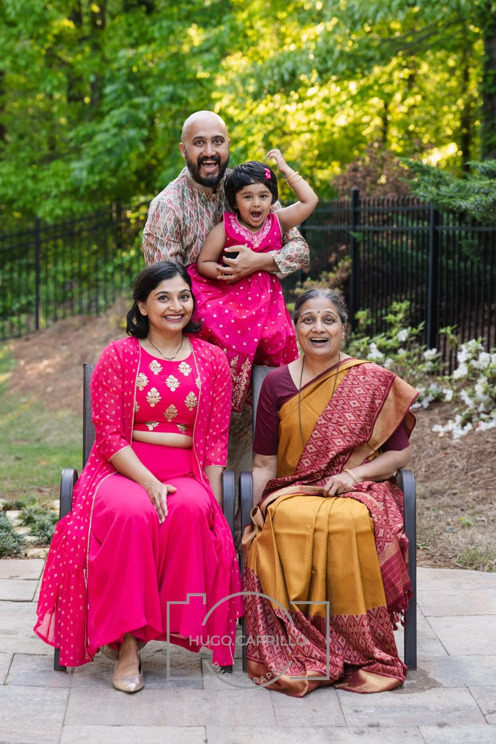 A multigenerational Indian family consisting of two women, a man, and a young girl, outdoors in a garden during daytime. They are smiling and dressed in colorful traditional Indian clothing, with green trees and a black fence in the background.