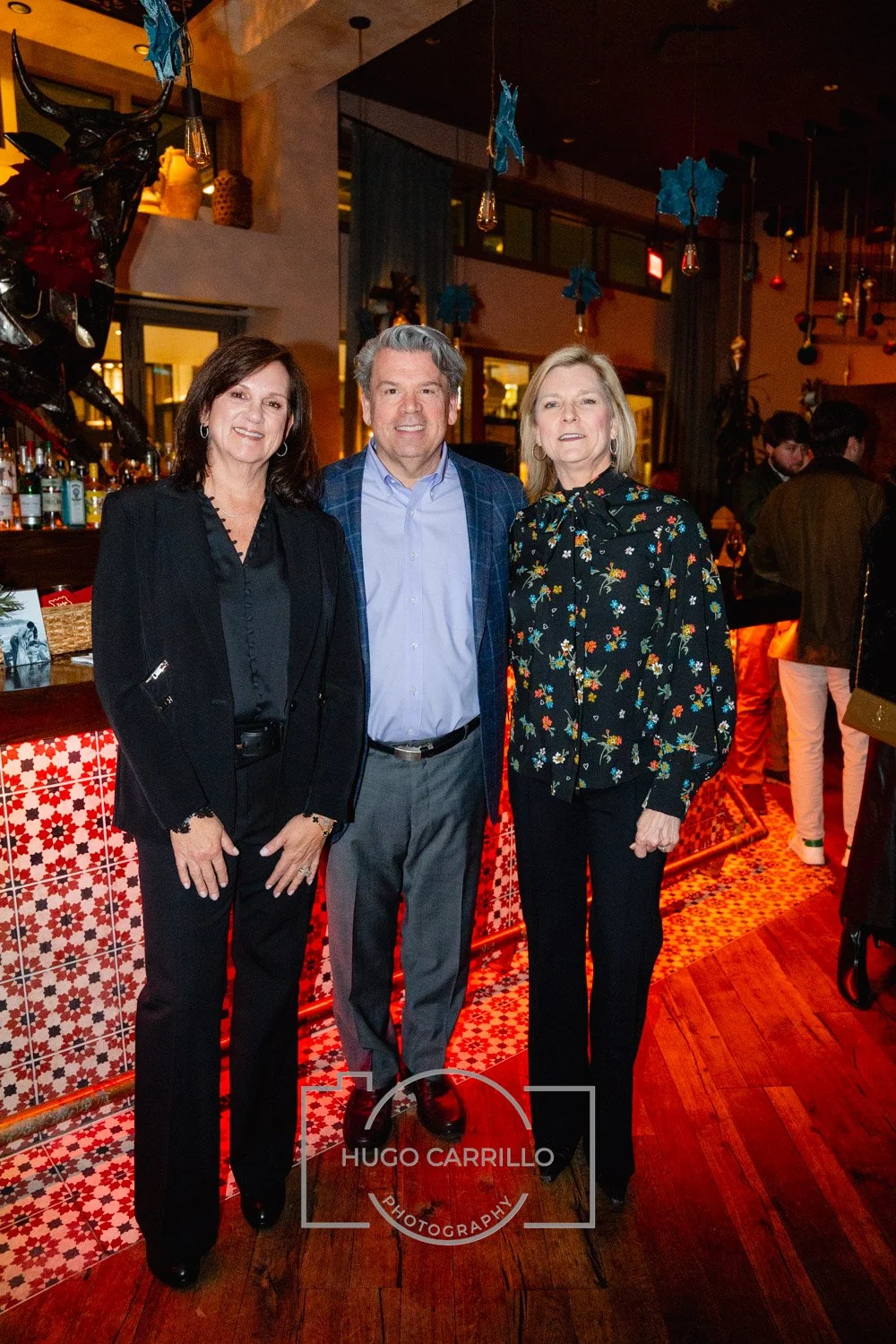 Three people standing together at a festive indoor event, dressed in formal attire, with a bar and Christmas decorations in the background.