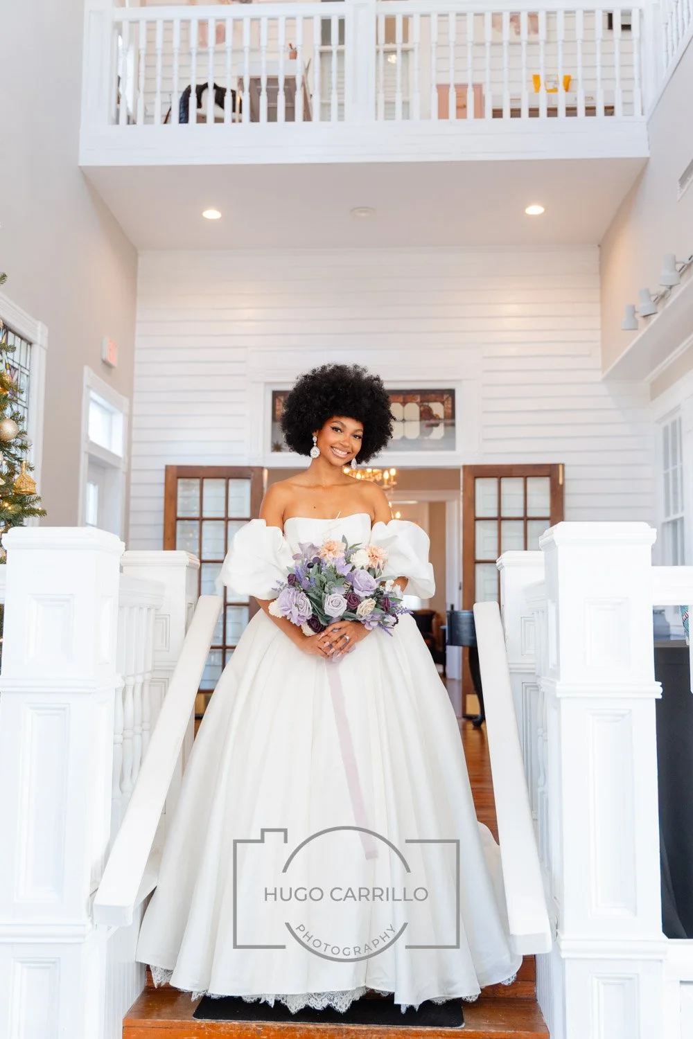 A smiling bride in a white wedding dress holding a purple and pink floral bouquet standing on a staircase inside a bright, white-walled house.