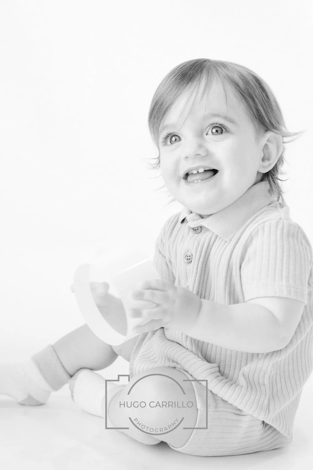 Child with light hair smiling and holding a cup, sitting on the floor in a striped outfit in a bright studio setting.