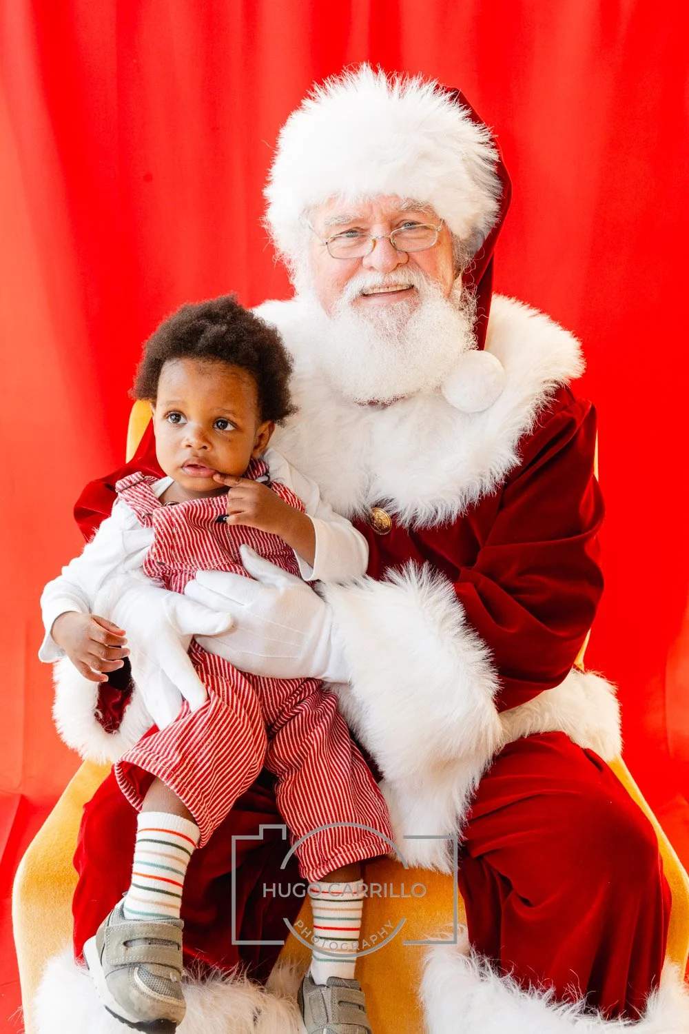 Santa Claus in a red suit with white fur trim holding a young child with curly hair, wearing a red and white striped outfit, against a red background.