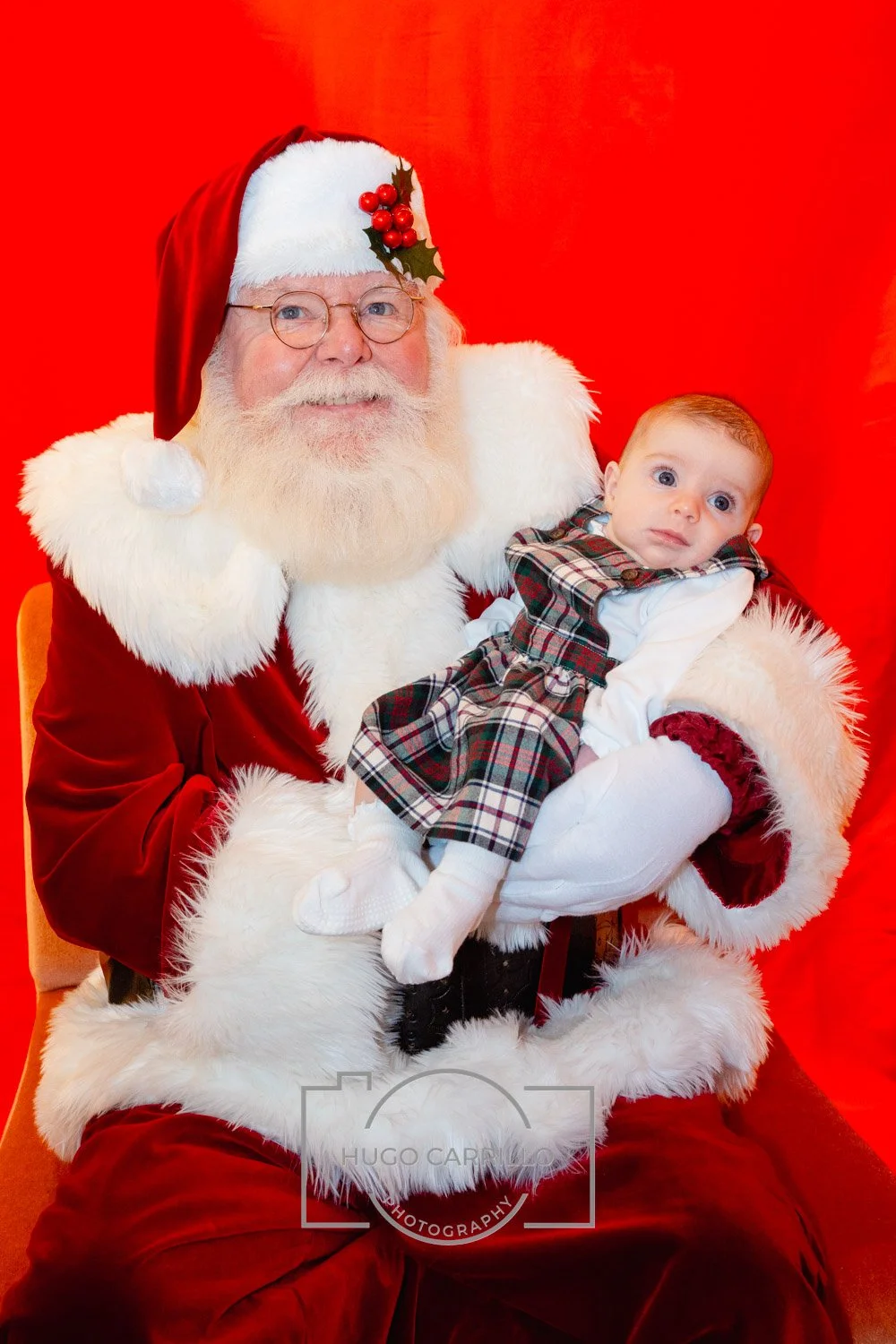 Santa Claus with glasses, a white beard, and a red hat with holly berries, holding a young child dressed in a plaid outfit against a red background.