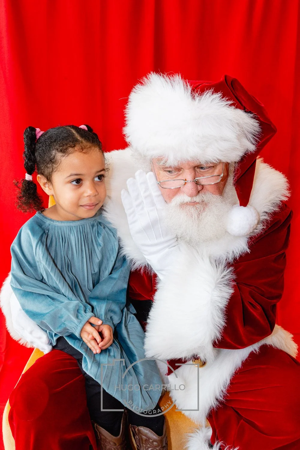 A young girl sitting on Santa Claus's lap, listening to him whisper while wearing a blue dress, with a red curtain in the background.