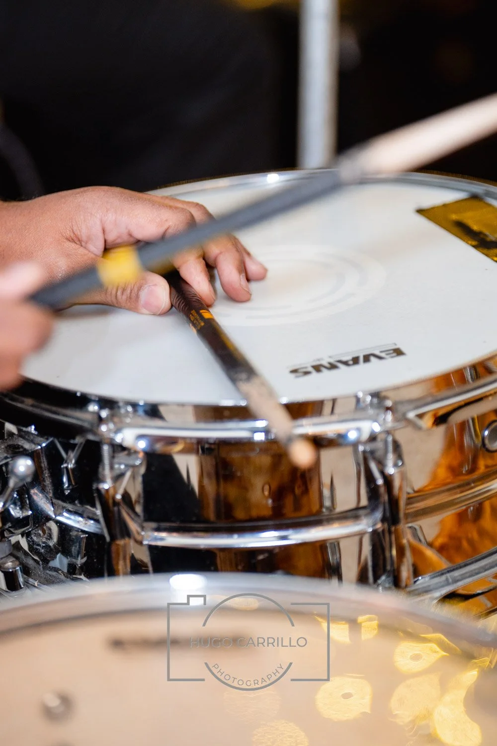 Close-up of a person's hands playing a snare drum with a drumstick, surrounded by other drums.