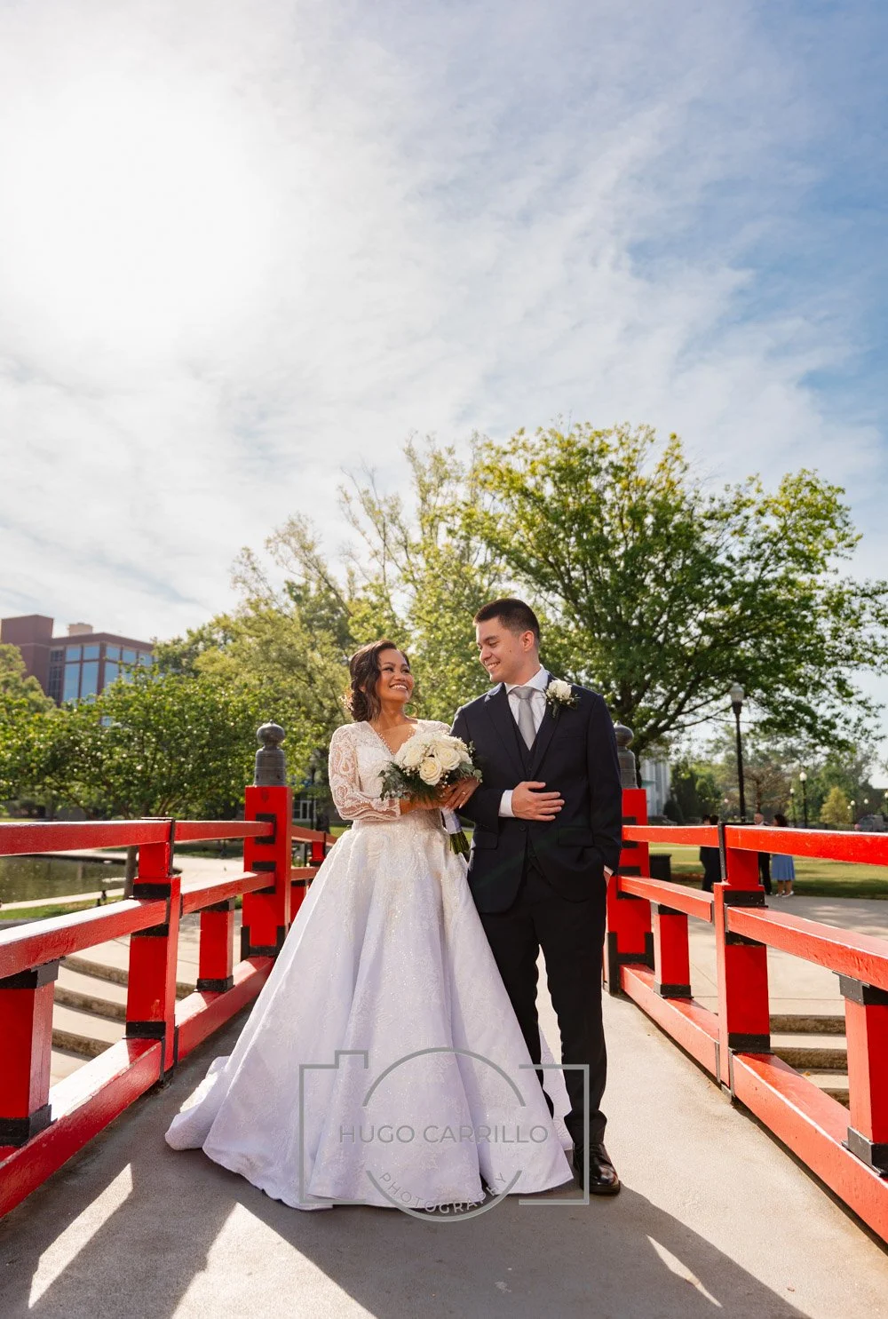 A bride and groom standing on a red bridge outdoors, smiling at each other. The bride is holding a bouquet of white flowers, wearing a white wedding dress, while the groom is wearing a dark suit and tie. There are green trees and a partly cloudy sky 