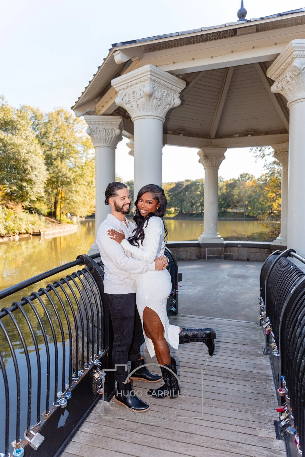 A couple hugging on a footbridge in a park, surrounded by trees with autumn foliage, near a river, with a sky in the background.