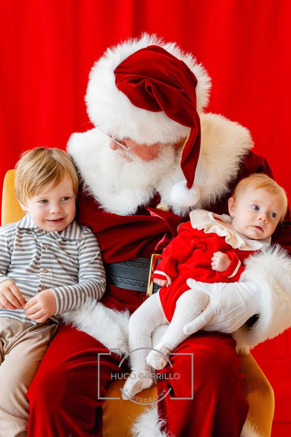 Santa Claus with two young children on his lap against a red background. The children are dressed in festive clothing, with one smiling and the other looking surprised.