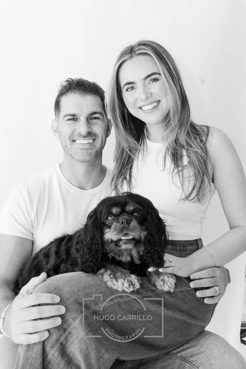 Black and white photo of a smiling man and woman posing with their dog, a Cavalier King Charles Spaniel, in front of a plain background.