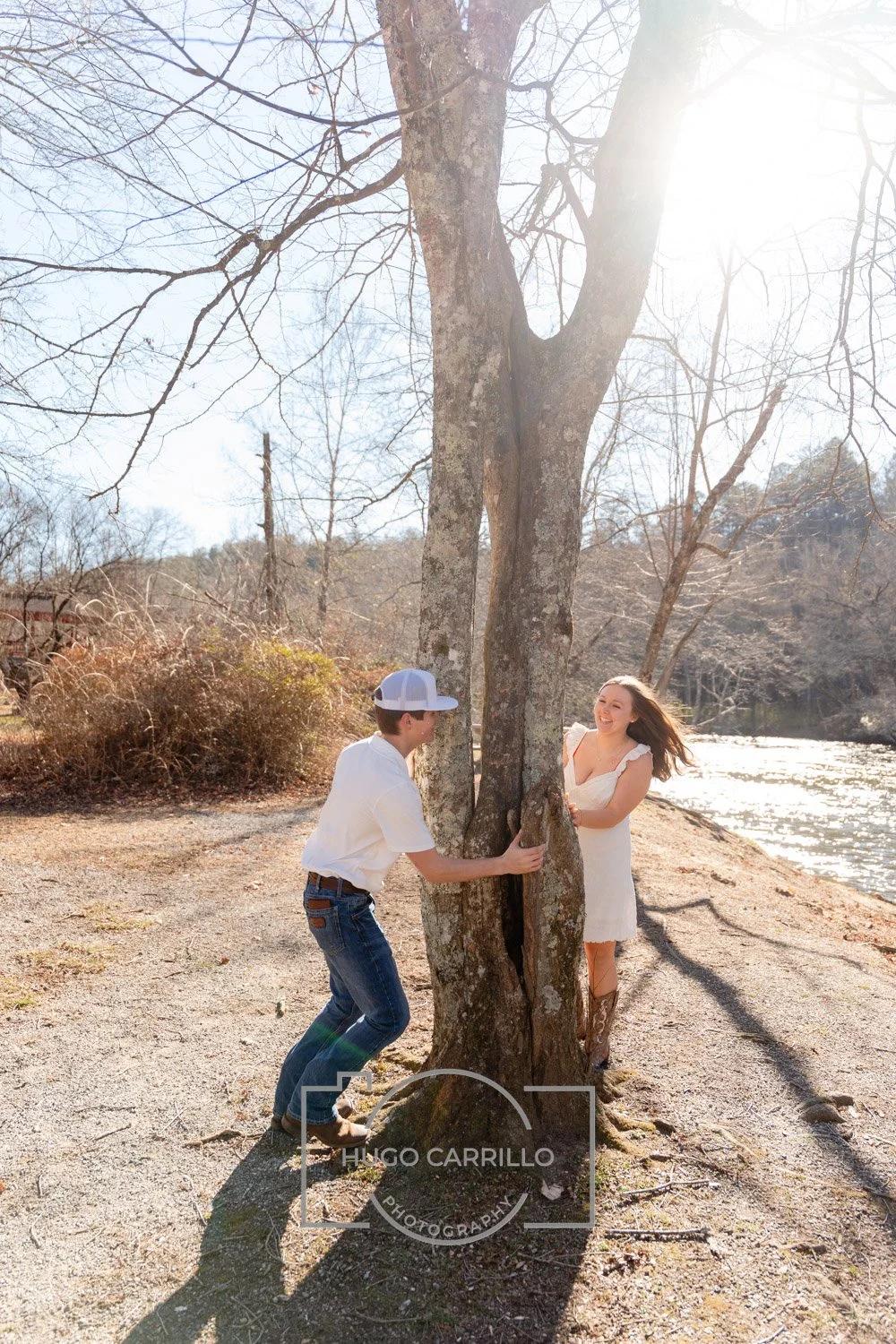 A young man and woman playfully hug around a large tree by a river, with sunlight filtering through the branches on a clear day.
