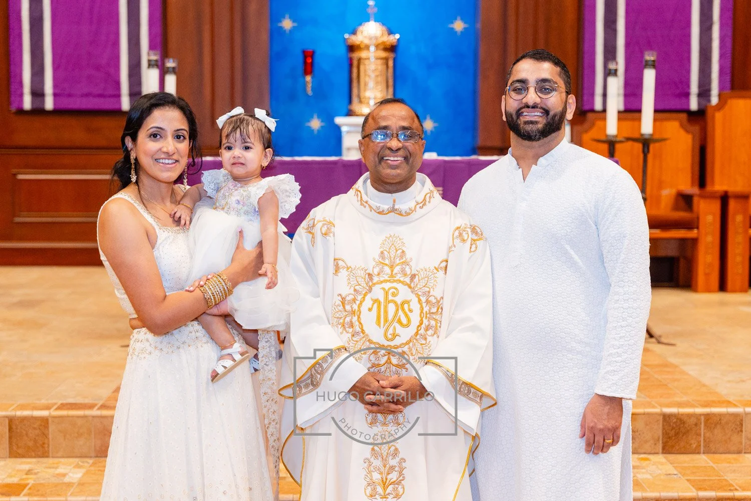 Family at a church ceremony, with a woman holding a young girl, a priest in the center, and a man on the right, all dressed in white and standing in front of an altar with purple and blue decorations.