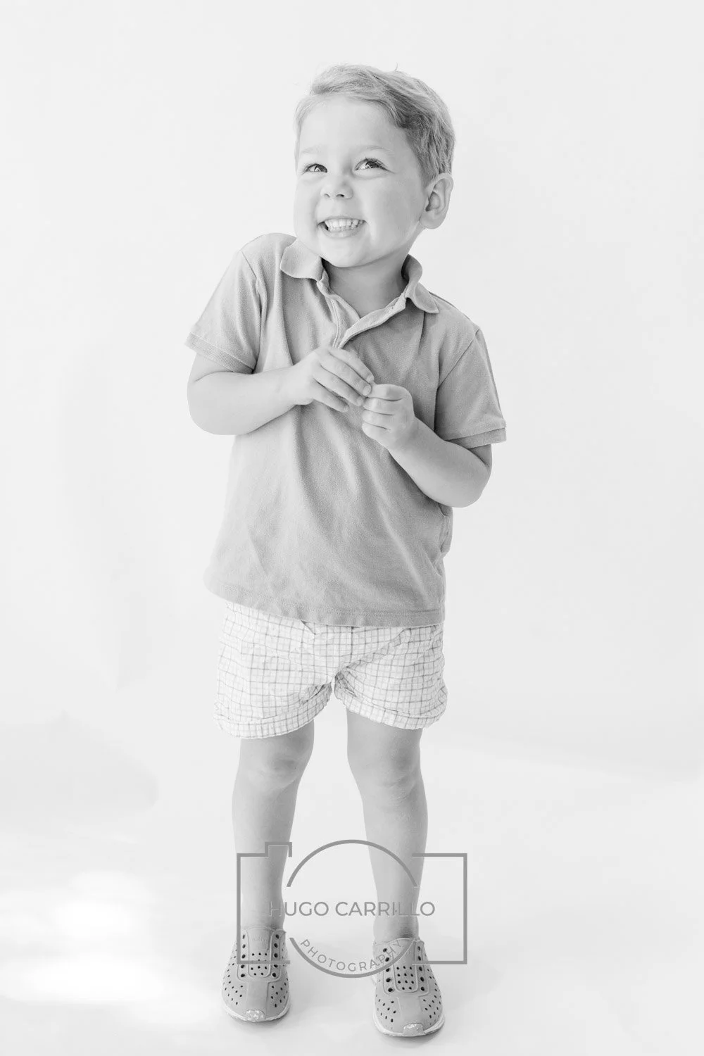 A young boy with short hair, smiling and looking upwards, wearing a polo shirt, checkered shorts, and sneakers, standing against a plain background.