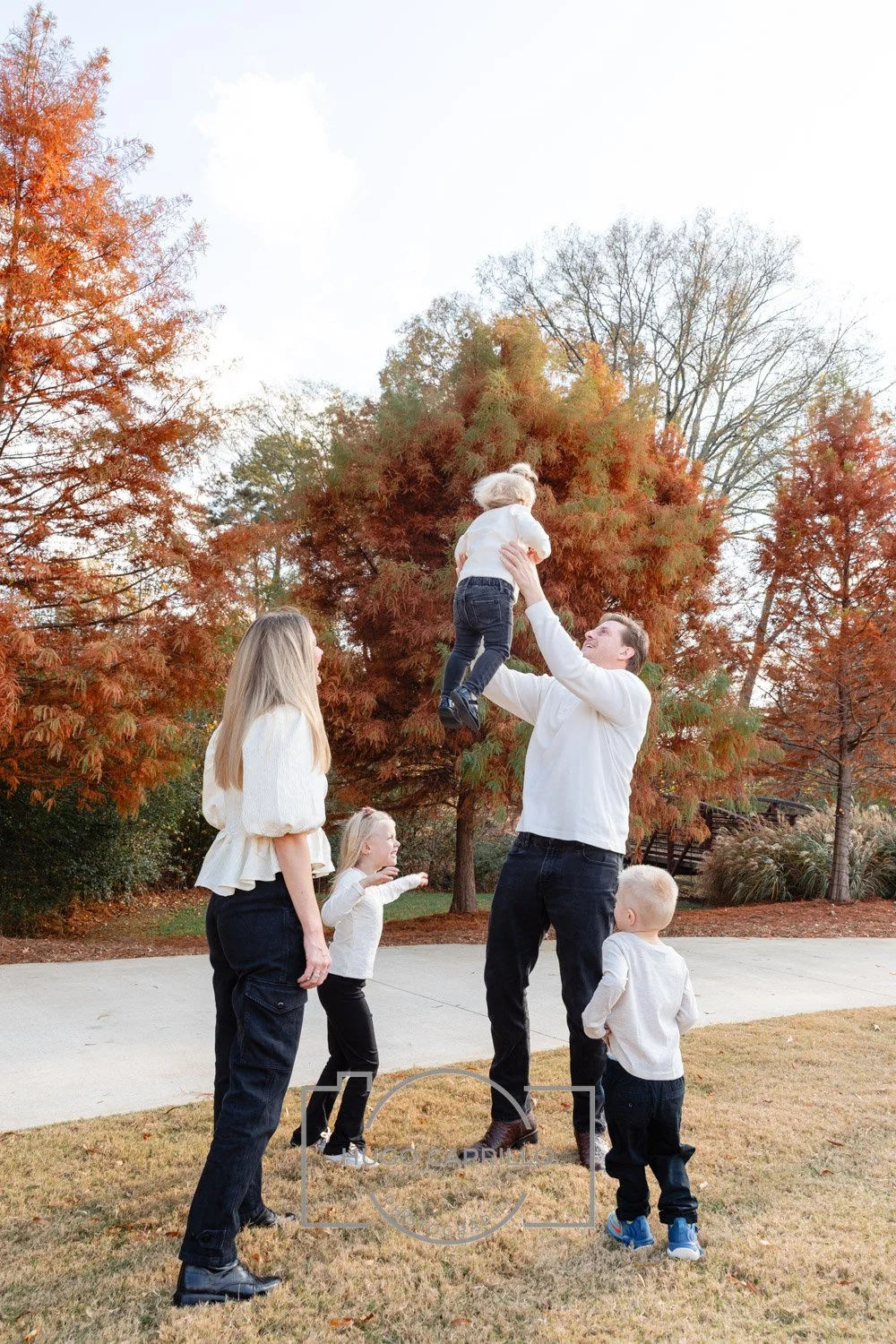 A family of five playing outdoors in autumn, with trees displaying fall foliage. A man lifts a young girl into the air while a woman and two children watch.
