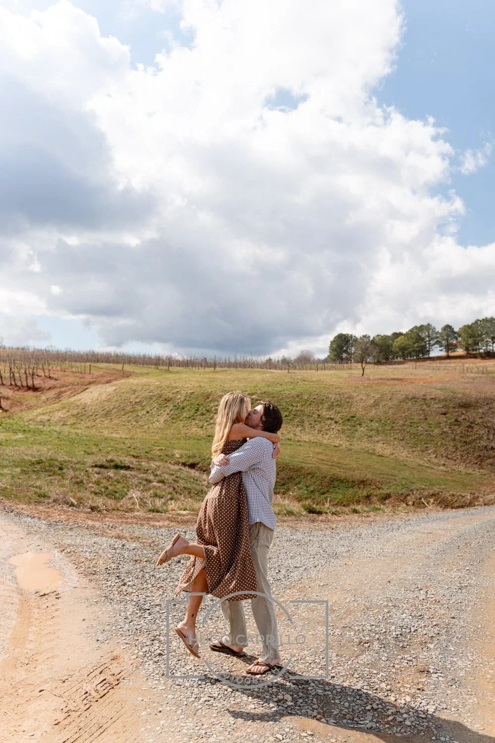 A couple is hugging and kissing on a gravel road in a rural outdoor setting with hills and trees under a partly cloudy sky.