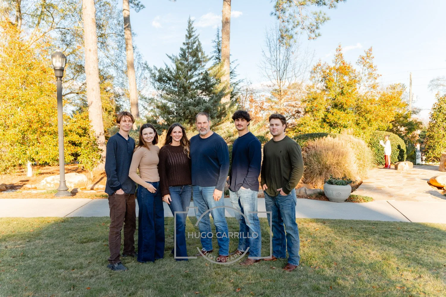 A group of six people standing on a lawn with trees and a pathway in the background during autumn. They are smiling and posing for the photo.