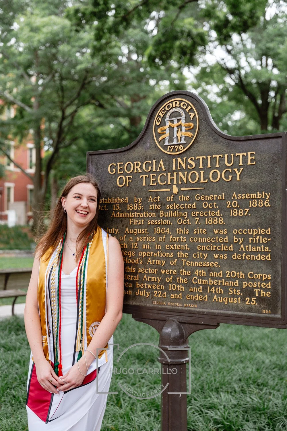 A young woman in a white dress and a gold graduation stole smiling and standing next to a historic marker sign at the Georgia Institute of Technology campus.