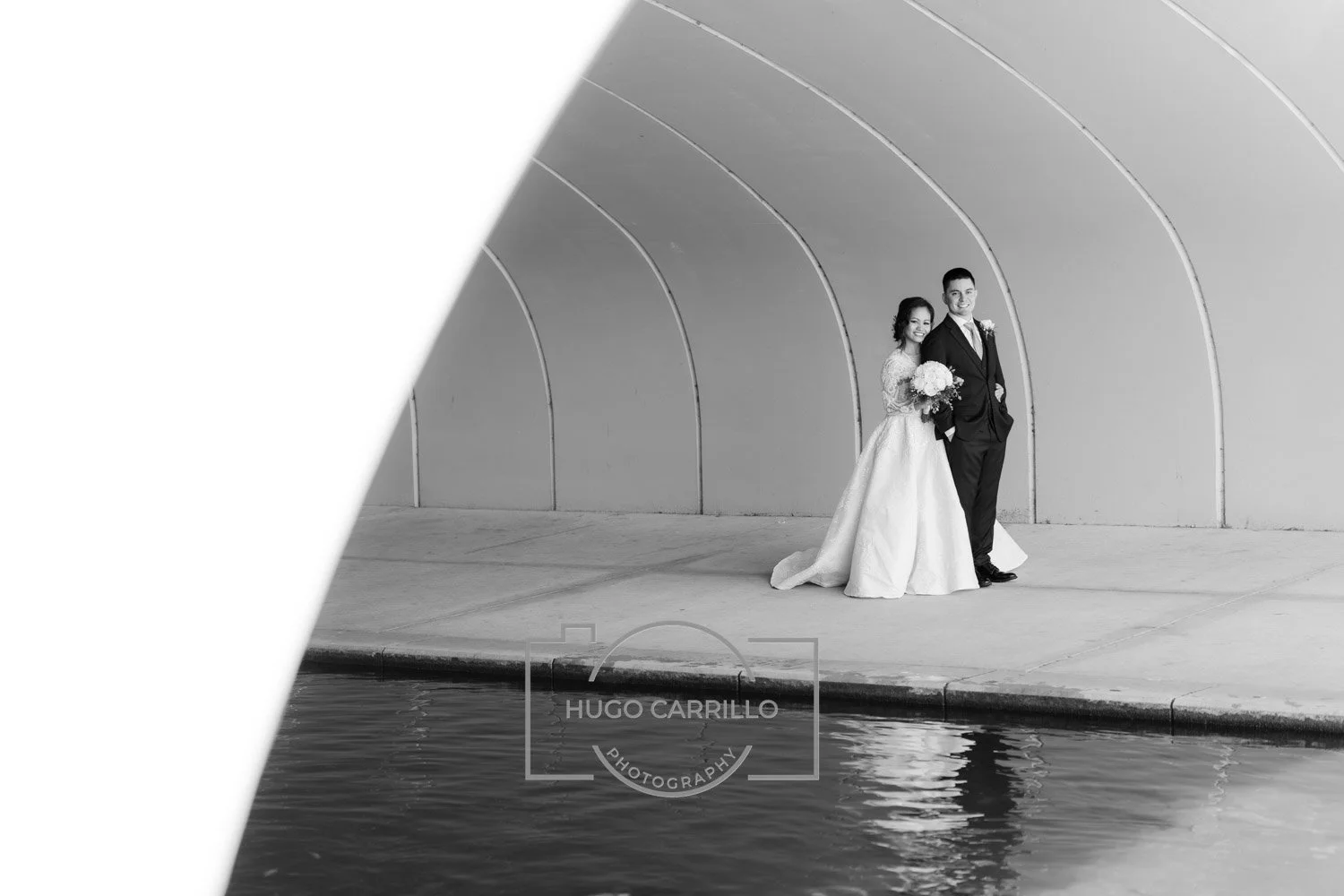 Black and white photo of a bride and groom standing close together on a sidewalk by water, with an arched wall in the background. The bride is holding a bouquet and wearing a long wedding dress, while the groom is in a suit. Their reflection is visib