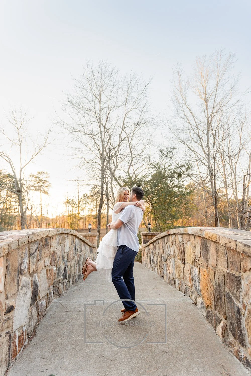 A couple dancing on a stone bridge with trees and a sunset in the background.