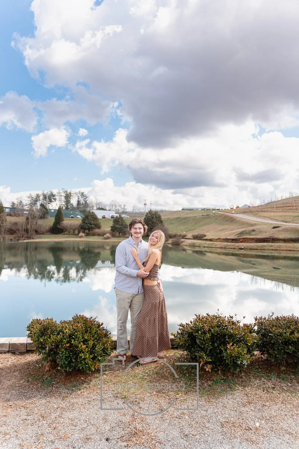 A couple stands by a pond, smiling and embracing each other with trees, a grassy hill, and a cloudy sky reflected in the water.
