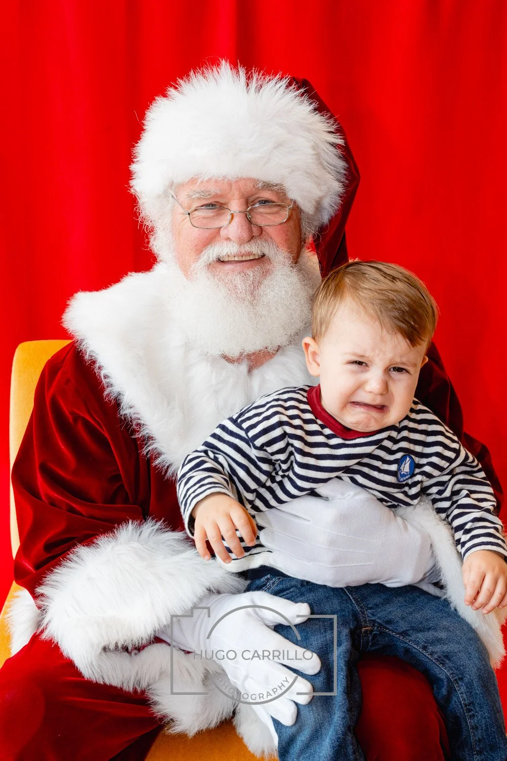 Santa Claus sitting with a young boy, who looks upset, against a red background.