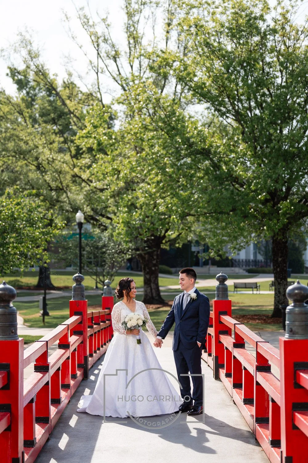 A bride and groom holding hands on a red bridge in a park with large green trees, sunlight, and a lamppost in the background.