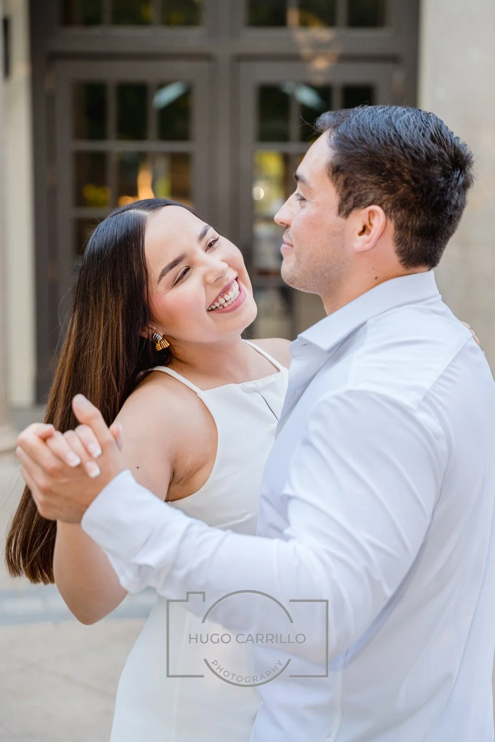 A couple dancing outdoors, smiling and looking happy, with the woman wearing a white dress and the man in a white shirt.