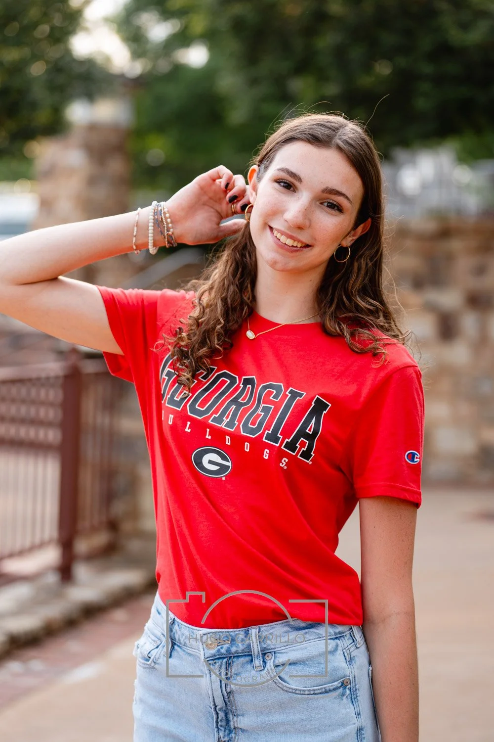 A young woman with long curly brown hair smiling outdoors, wearing a red Georgia Bulldogs T-shirt and light blue jeans, with a bracelet and necklace, standing near a fence with trees in the background.
