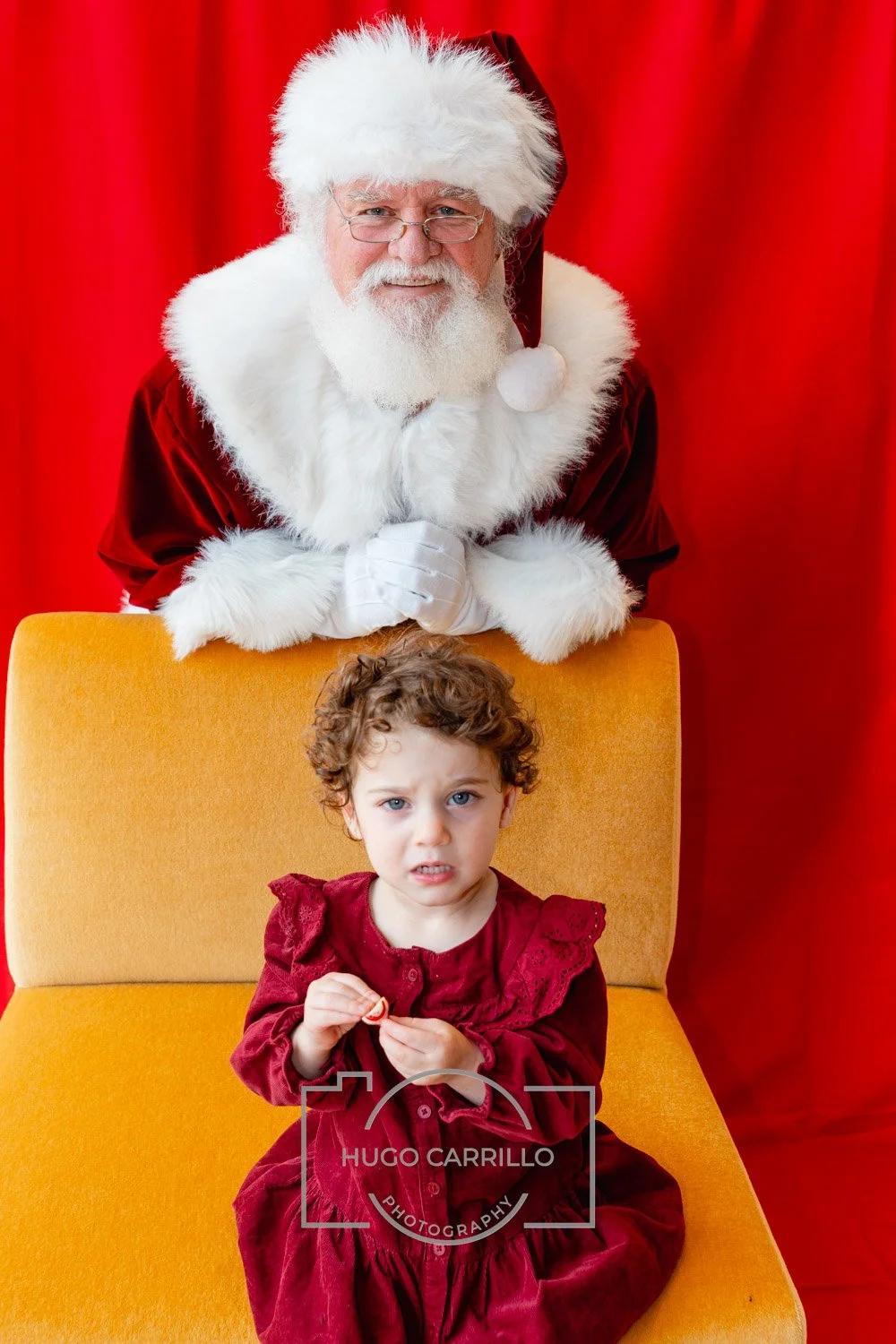 Santa Claus with glasses and a white beard leaning over a yellow chair behind a young girl with curly hair and a red dress, on a red background.