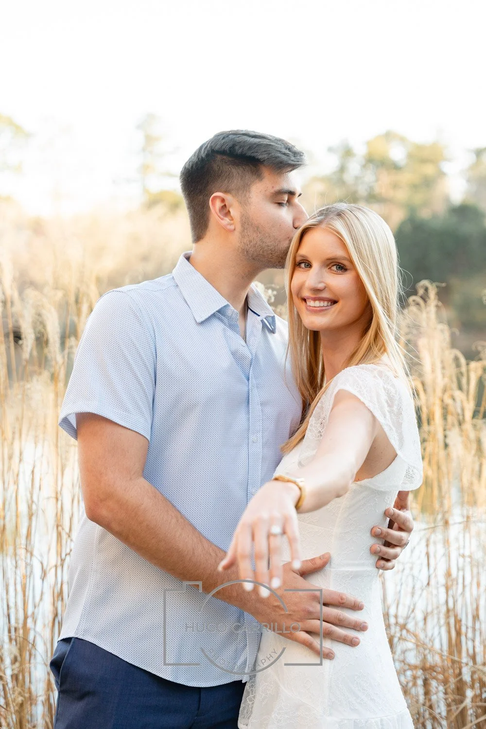 A young couple stands outdoors in a natural setting with tall grass and trees in the background. The man is kissing the woman on the forehead while she smiles at the camera, extending her arm to display a ring on her finger.