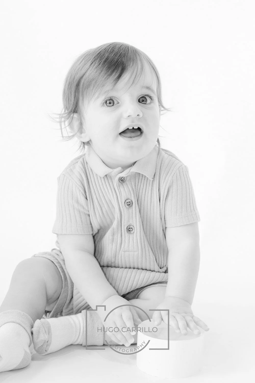Black and white photo of a young child sitting on the floor, with one leg bent and the other extended, wearing a short-sleeved collar shirt and shorts. The child has short hair and an expressive face with an open mouth, looking slightly upward.