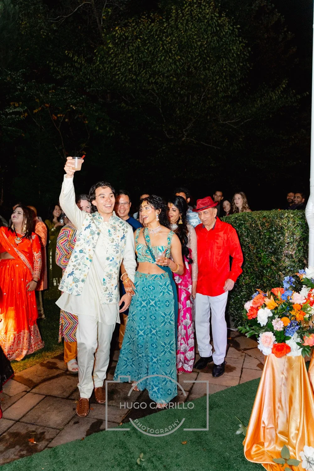 People celebrating at a wedding or festive event, with a man in a floral vest raising a toast and women in colorful dresses smiling, outdoor at night with flowers and greenery.