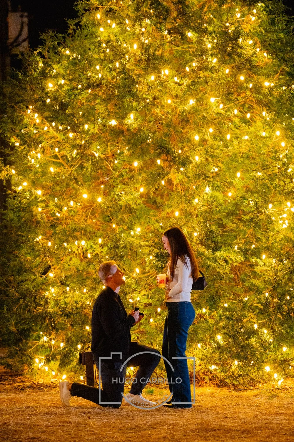 A man proposes marriage to a woman at night in front of a large decorated Christmas tree with lights. The man is kneeling holding a ring box, while the woman is smiling and holding a drink, with a black bag on her shoulder.
