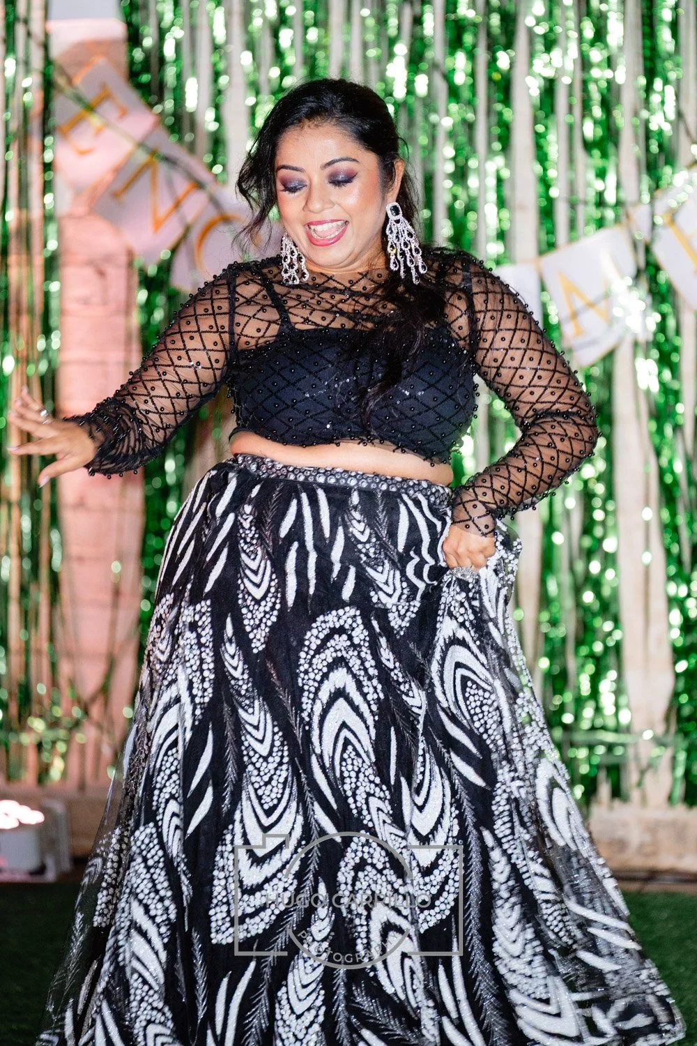Woman wearing a black mesh top with long sleeves and a black bralette, paired with a black and white patterned skirt, dancing at a celebration with green and gold decorations in the background.