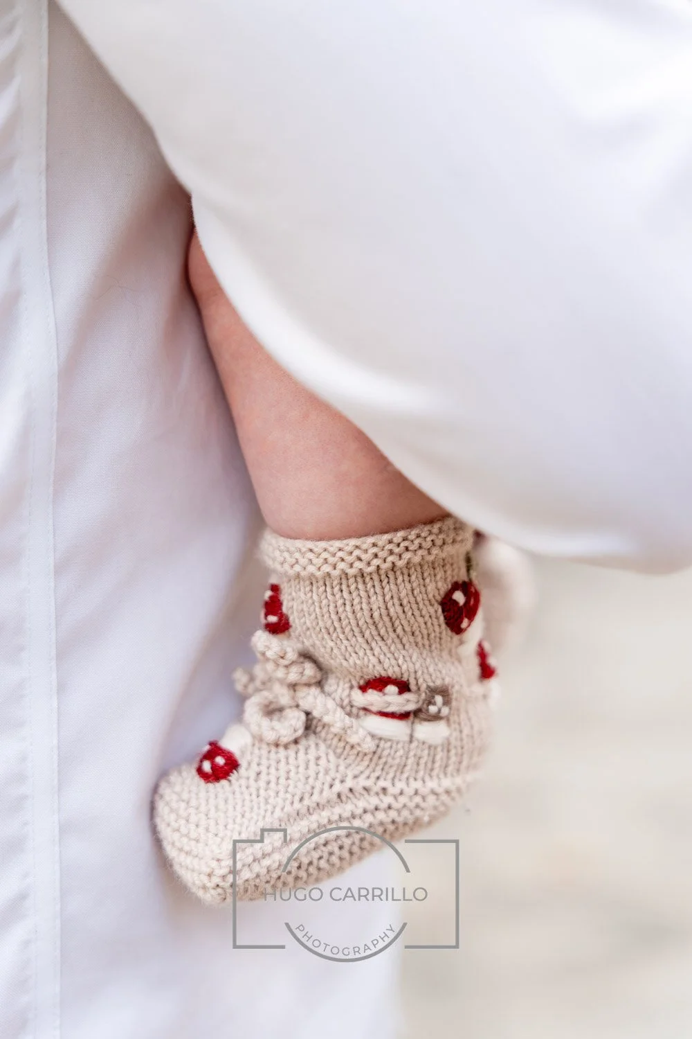Close-up of a child's ankle with a beige knitted sock featuring red and white mushroom patterns, partly covered by a white coat or shirt.
