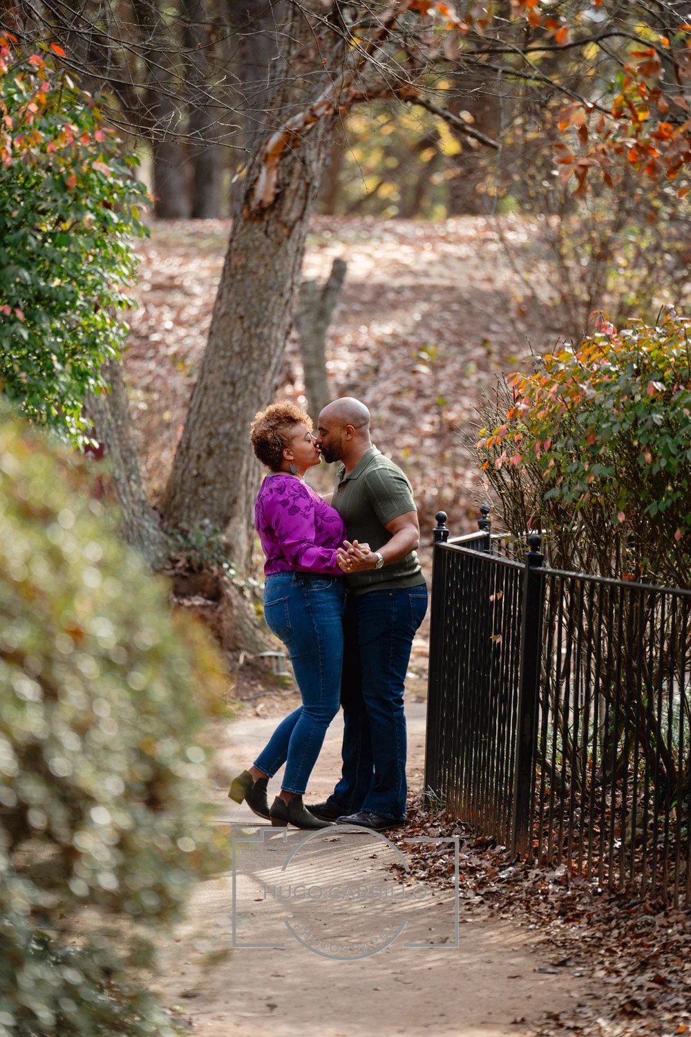 A couple standing very close, holding hands, and about to kiss in a wooded park during autumn with fallen leaves on the ground.