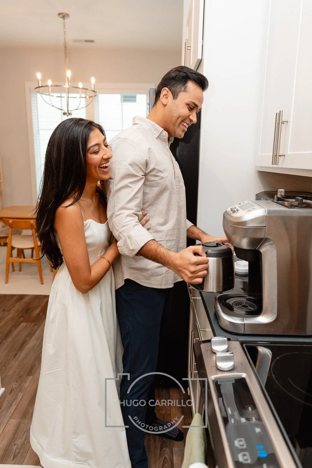 A young woman in a white dress stands behind a man as he makes coffee in a kitchen. They are both smiling and appear happy.