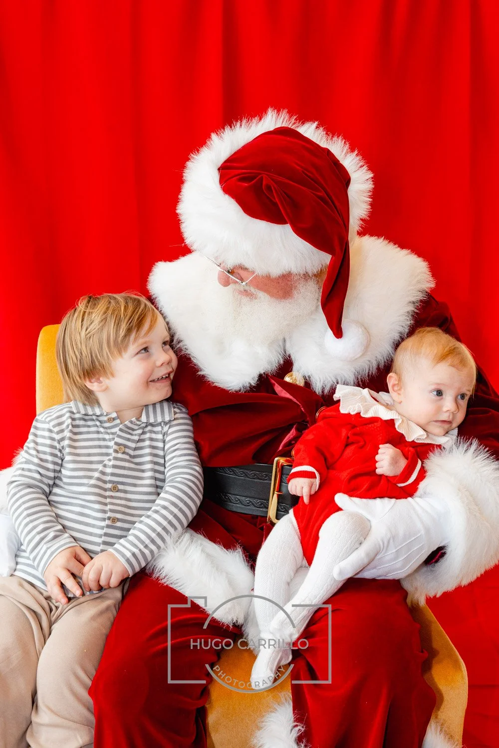 Santa Claus sitting with two young children, one boy and one girl, against a red curtain background. Santa is holding the girl, who is wearing a red dress, and the boy is sitting beside Santa, smiling happily.