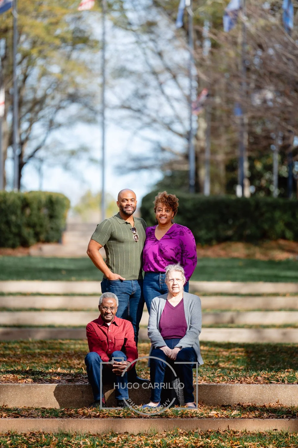 A group of five diverse adults posing outdoors on steps in a park with trees and flags in the background.