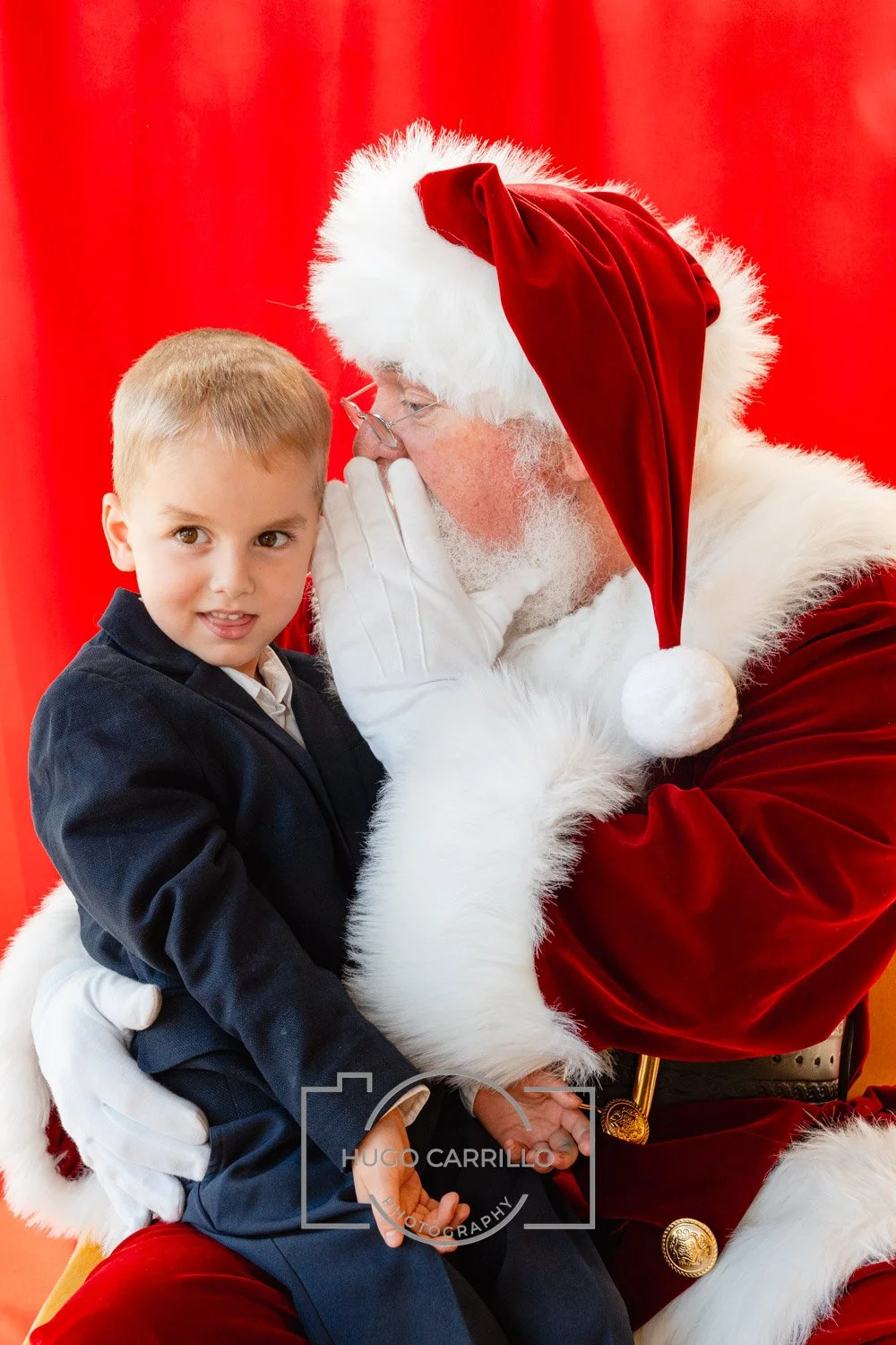 A young boy sitting on Santa Claus's lap, whispering to Santa, who is dressed in a traditional red and white Santa outfit with a red hat, white beard, and glasses, against a red background.