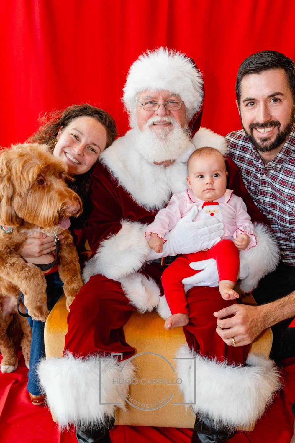 A family with their dog taking a Christmas photo with Santa Claus, who is dressed in a red suit with white fur trim, and a baby girl in red pajamas with a snowman on the front, all sitting in front of a red curtain.