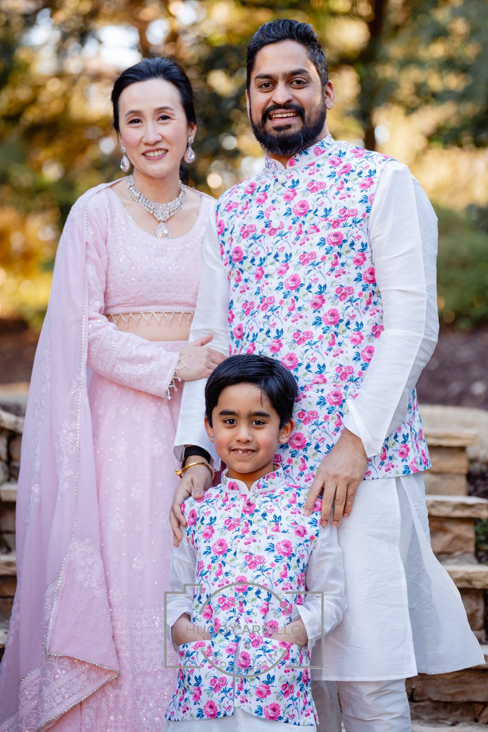 A family of three posing outdoors during fall, dressed in traditional Indian attire: the woman in a pink saree, the man in a white kurta with a floral vest, and the boy in a matching floral kurta.