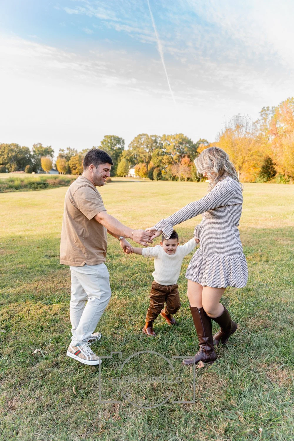 A family of three playing outdoors in a grassy park during autumn, holding hands and swinging a young boy between them.