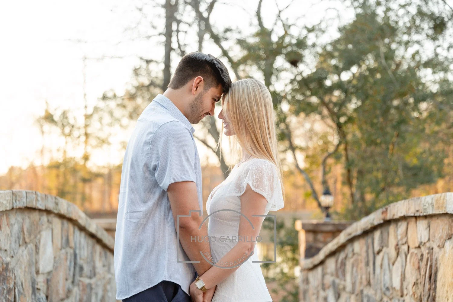 A man and woman standing close on a stone bridge at sunset, with their foreheads touching and holding hands, outdoors with trees in the background.