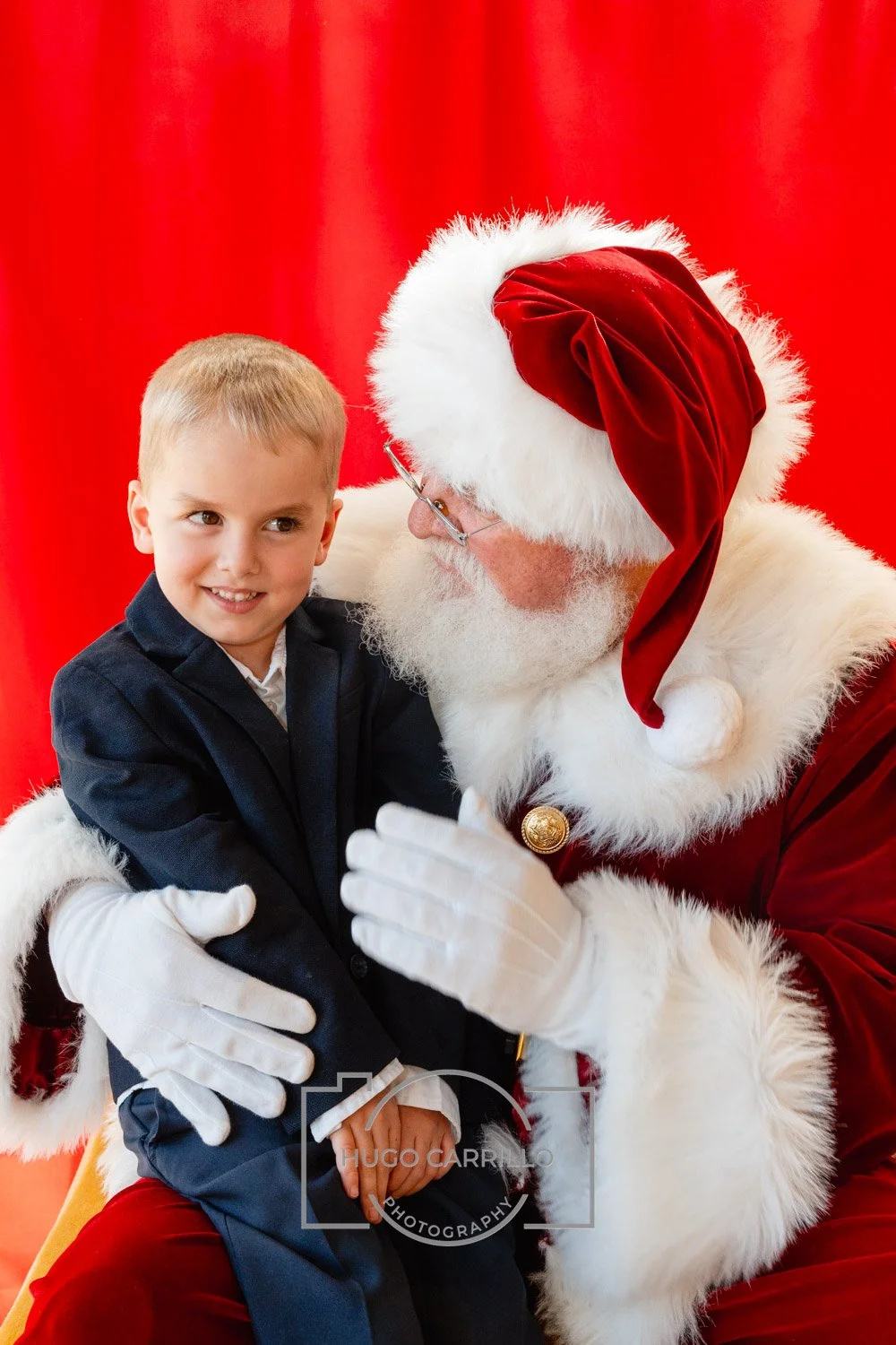A young boy in a dark suit sitting on Santa Claus's lap, who is dressed in a traditional red Santa outfit with a white fur trim, against a red curtain background.