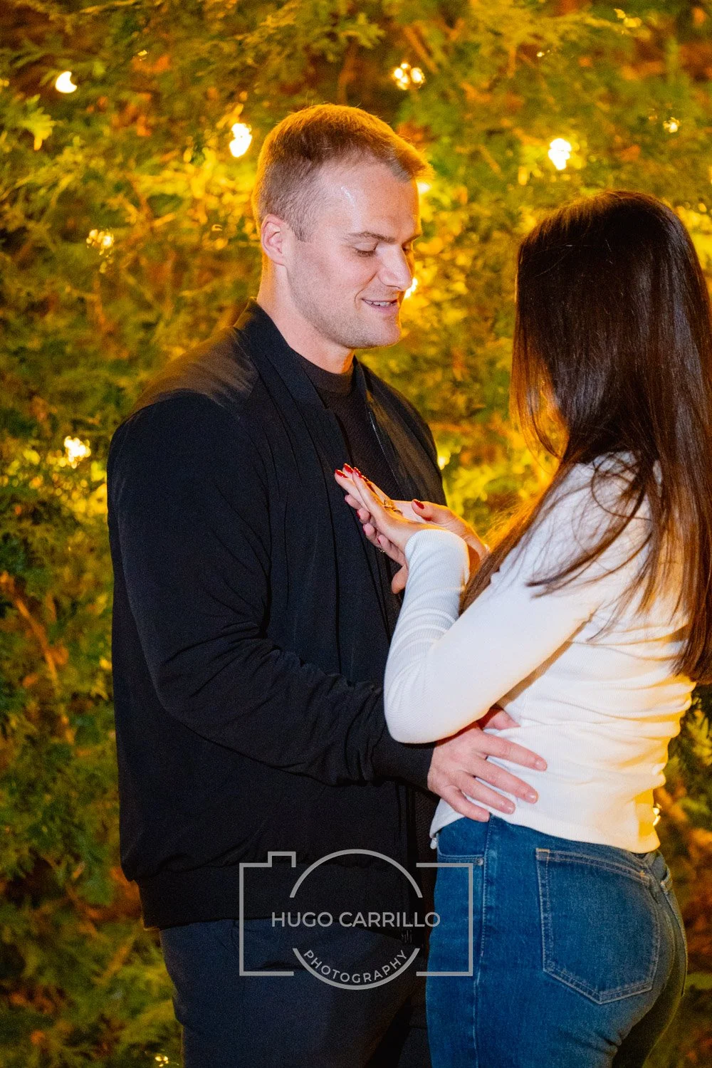 A couple standing close together outdoors at night, with warm yellow lights and trees with autumn leaves in the background. The woman is touching the man's chest, and they are smiling at each other.