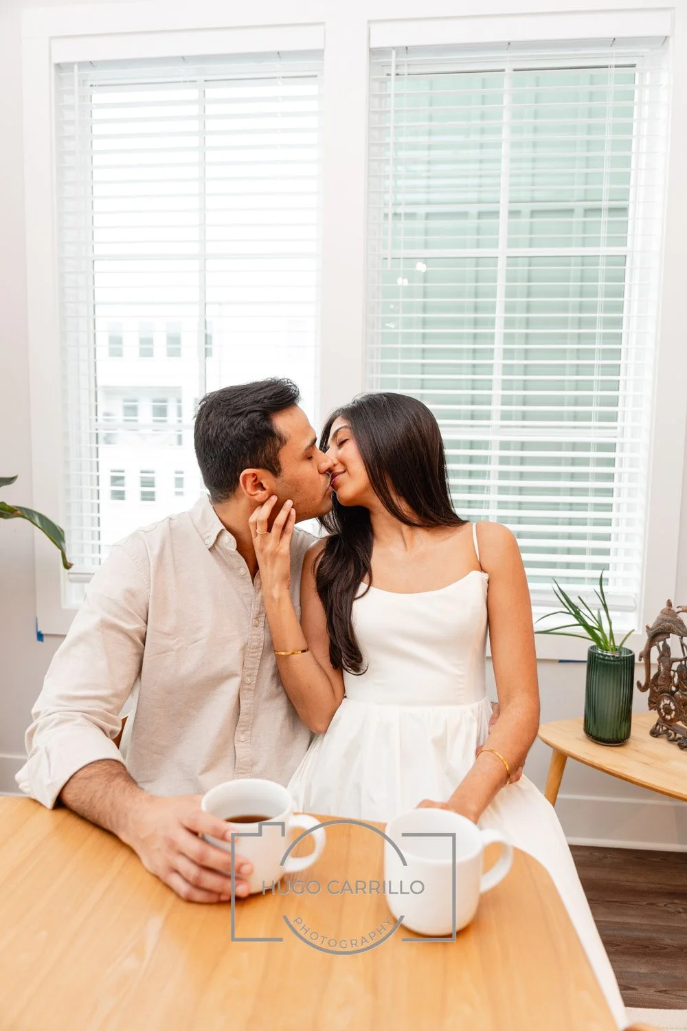 A couple sharing a romantic kiss at a kitchen table with coffee mugs, in a bright room with large windows and white blinds.