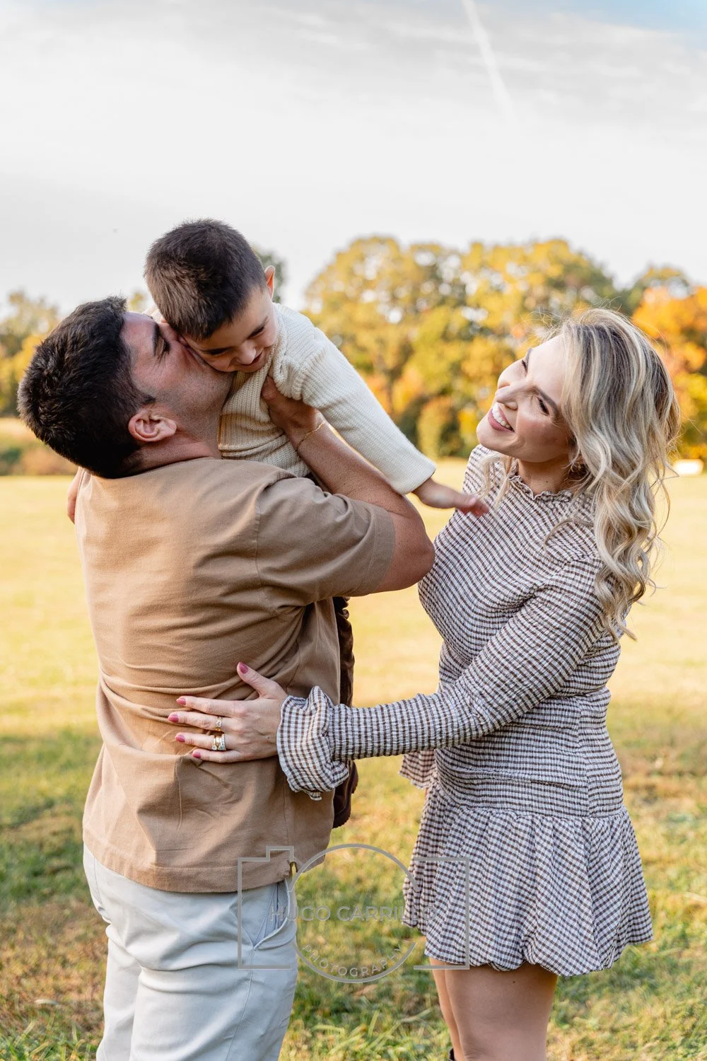 A happy family of three enjoying a moment outdoors in a park during autumn. The father lifts his young son upward, and the mother smiles at them, all amid fall foliage.